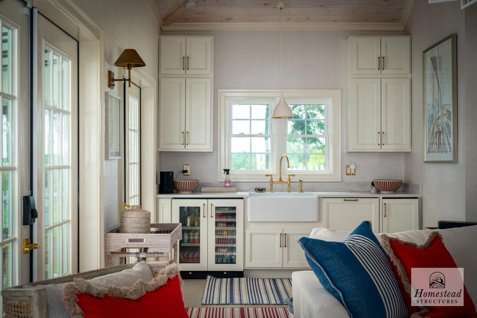 Cozy kitchen with white cabinets, a large farmhouse sink, and a window overlooking greenery. Decor includes striped pillows, artwork, and a small bar cart.