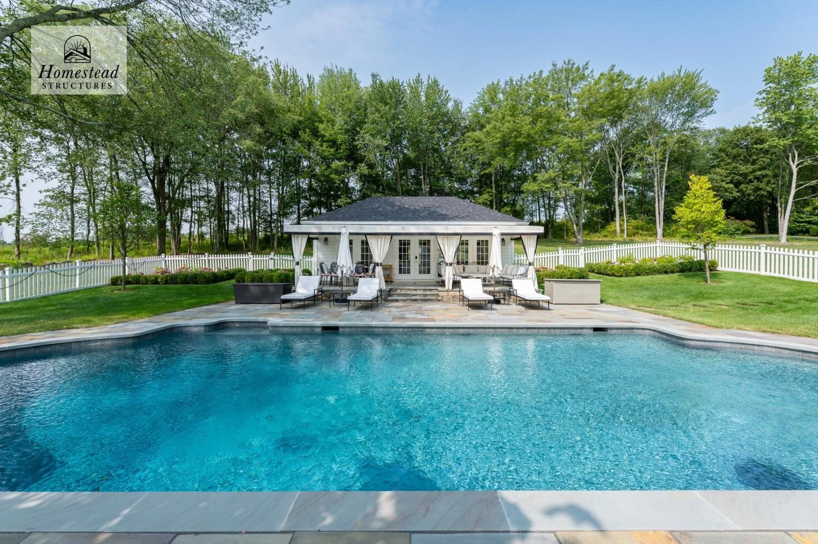 Backyard with swimming pool, poolside lounge chairs and umbrellas, patio area with outdoor furniture, small trees, green grass, and a white fence surrounding the yard. A house with a dark roof and white exterior is visible in the background, with a w