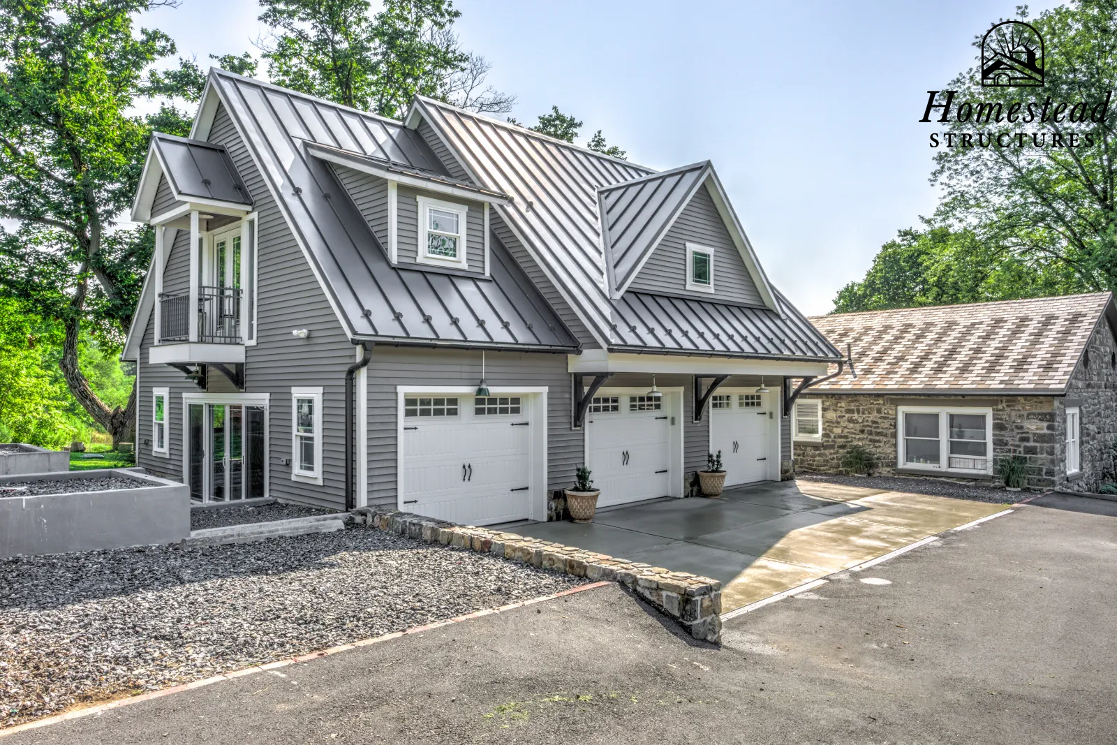 New gray house with metal roof, multiple garage doors, small balcony, and a stone section, surrounded by trees.