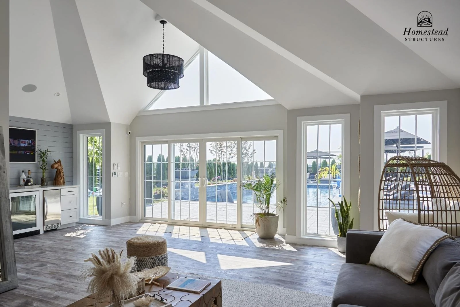 Bright living room with large glass sliding doors leading to a pool area, featuring a gray sofa with a white pillow, a hanging rattan chair, plants, and light wood flooring, with sunlight streaming in.