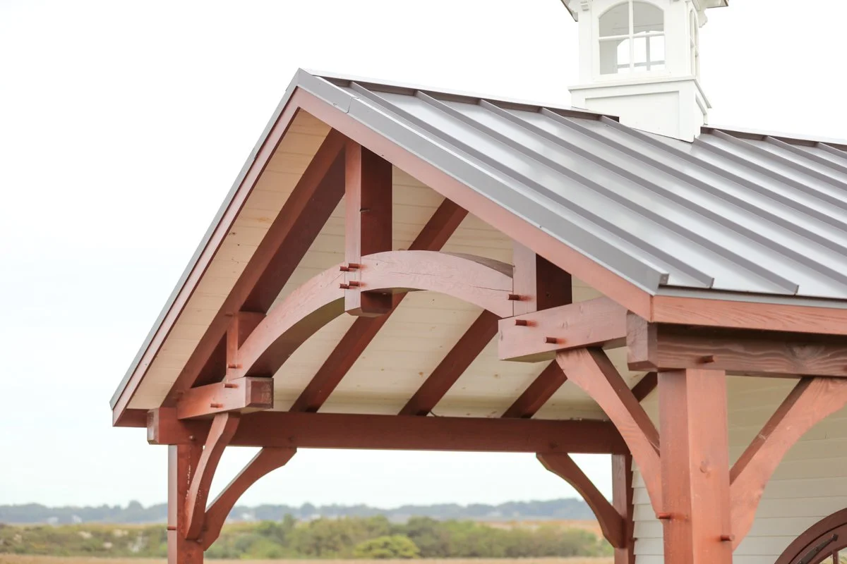 Close-up of a wooden porch with a metal roof and decorative support beams, overlooking a landscape with trees in the distance.