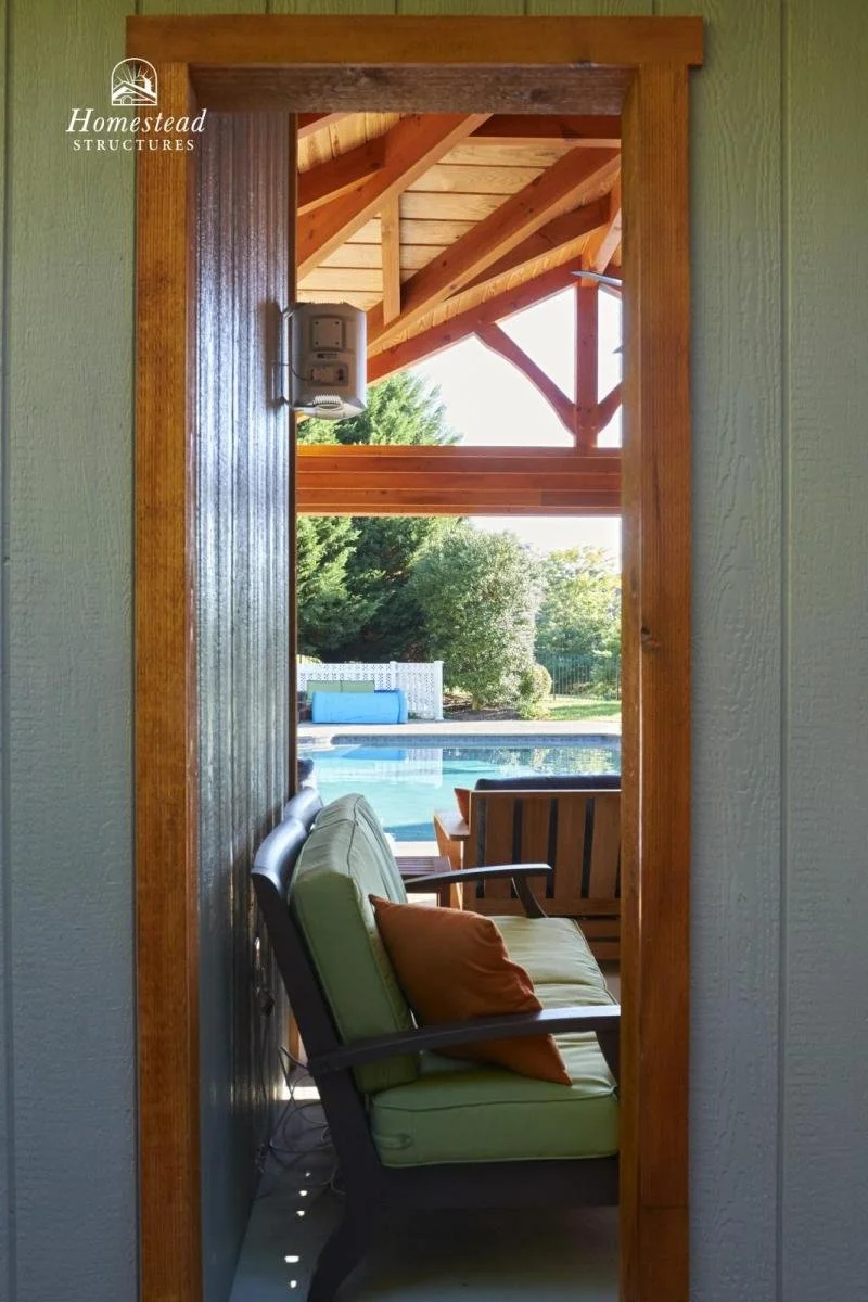A view through a wooden door frame showing an outdoor seating area with a swimming pool, wooden chairs, and lush green trees in the background.