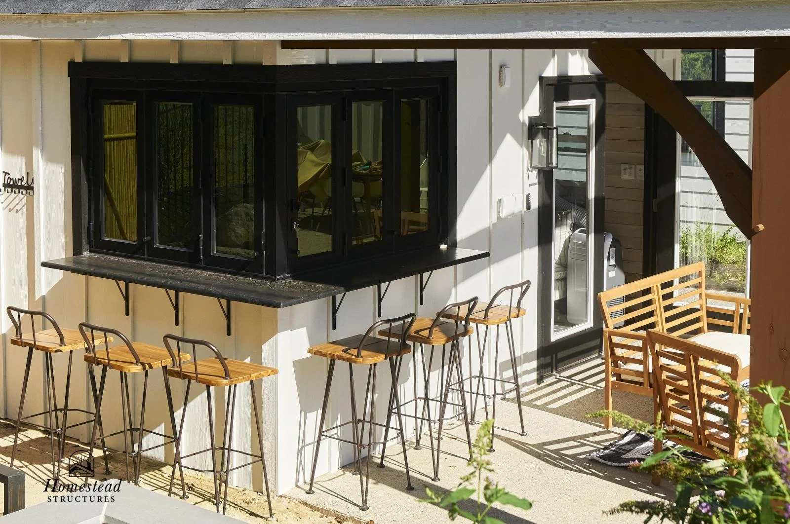 Exterior patio with six wooden bar stools with metal frames lined up along a black stone countertop beneath a balcony, with a wooden seating area and potted plants in the background.