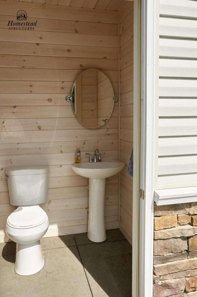 An outdoor bathroom with a toilet, a pedestal sink, a mirror, and a wooden wall. The bathroom appears to be part of a portable or small structure. There is a soap dispenser on the sink and a towel hanging on the wall.