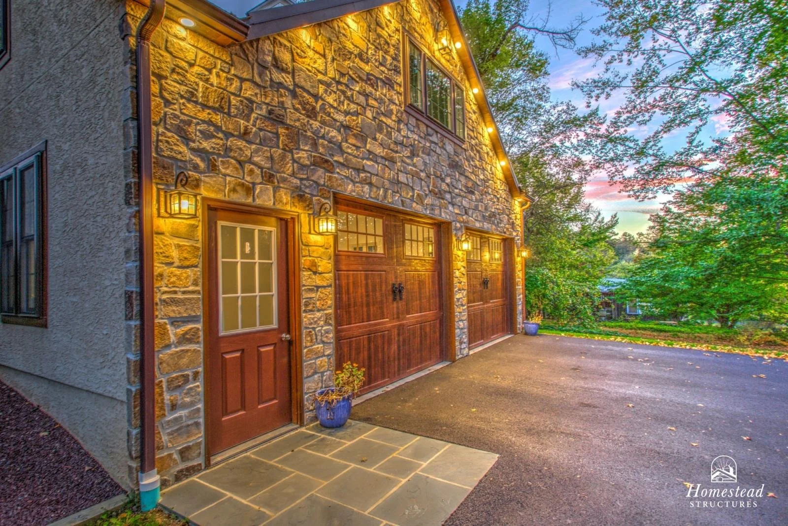 Exterior view of a house with a stone facade, wooden garage doors, and a wooden door with glass panels, illuminated by warm outdoor lighting at dusk. The driveway is paved, and there are trees and a colorful sky in the background.