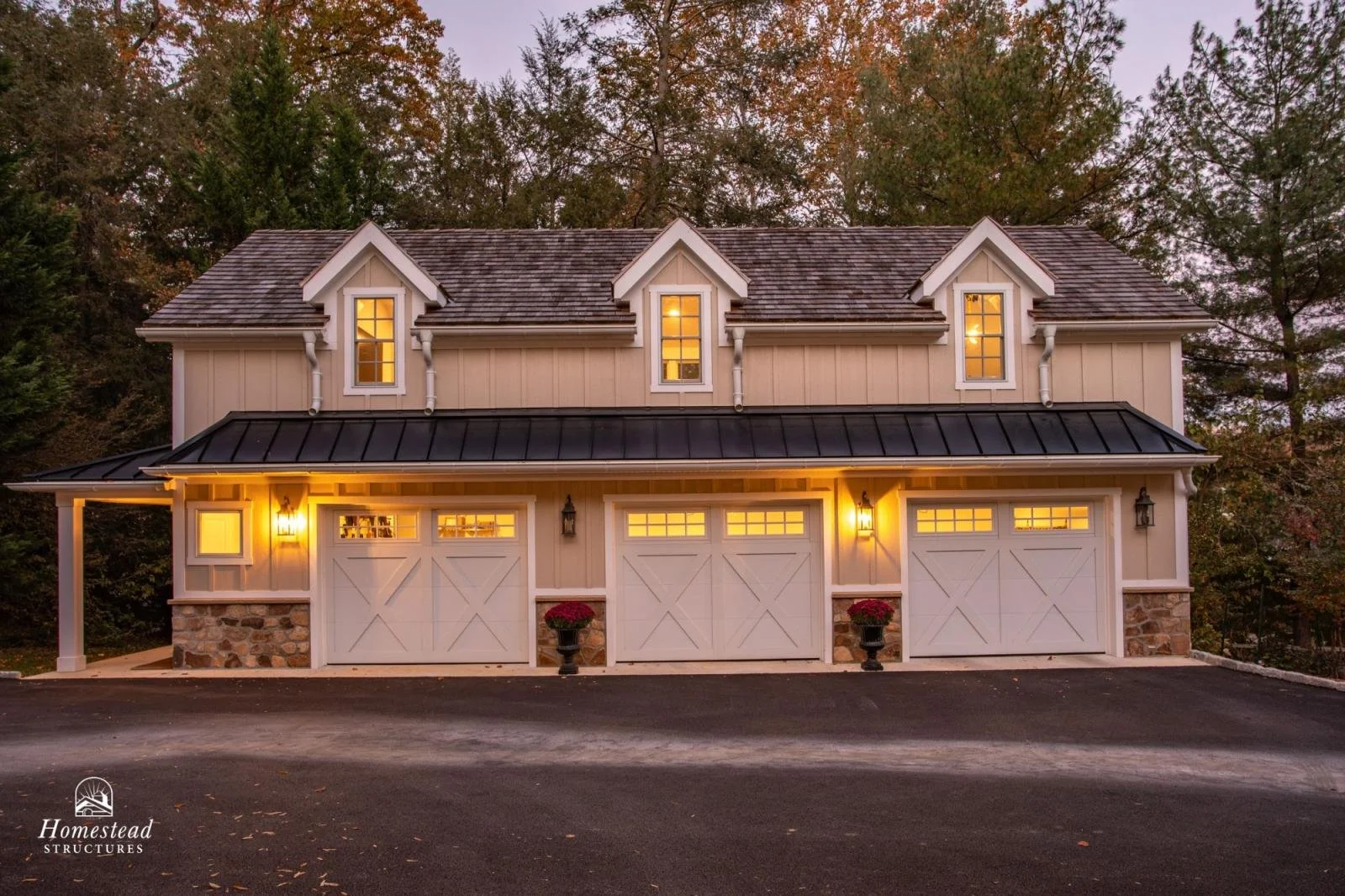A two-story garage with three white doors, stone foundation, and warm exterior lighting, surrounded by trees at dusk.