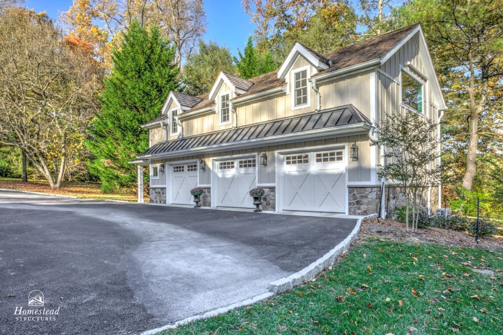 A two-story garage with three white doors, stone bases, and black metal roofing, surrounded by trees with fall foliage.