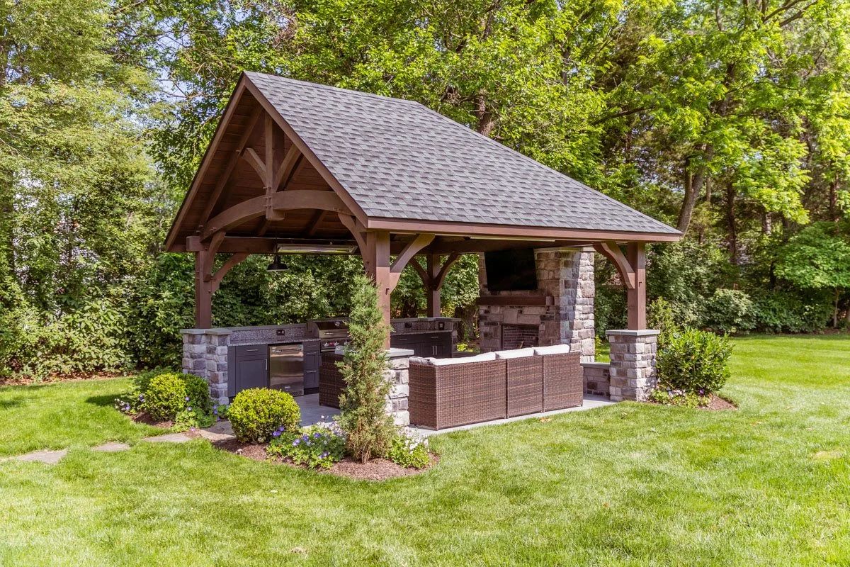 An outdoor pavilion with a shingle roof, stone columns, built-in grill, and a mounted TV, surrounded by a well-maintained lawn and lush trees.