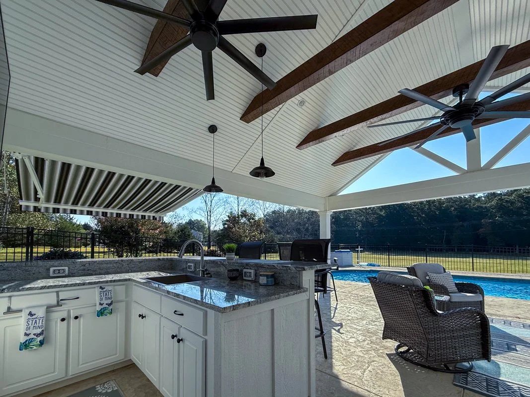 Outdoor covered patio with kitchen area, stainless steel sink, granite countertop, black pendant lights, ceiling fans, wicker chairs, swimming pool, and a spacious backyard with trees and a fence in the background.