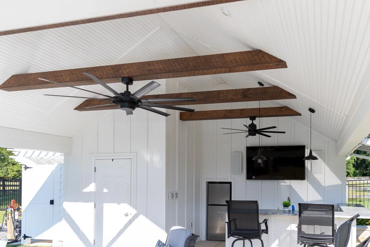 Outdoor patio area with white wooden walls and ceiling, exposed wooden beams, ceiling fans, a mounted TV, small refrigerator, and black patio chairs.
