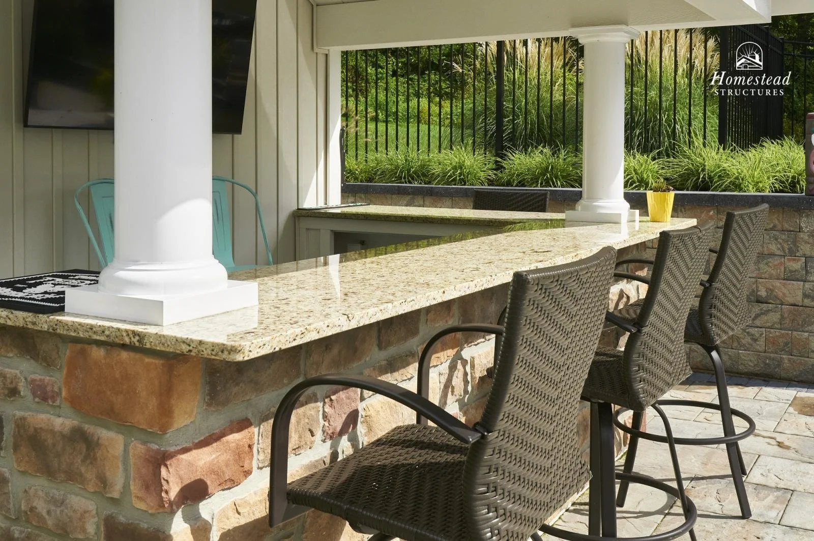 Outdoor bar area with a granite countertop, wicker bar stools, a yellow potted plant, and a black metal fence with green plants behind it.