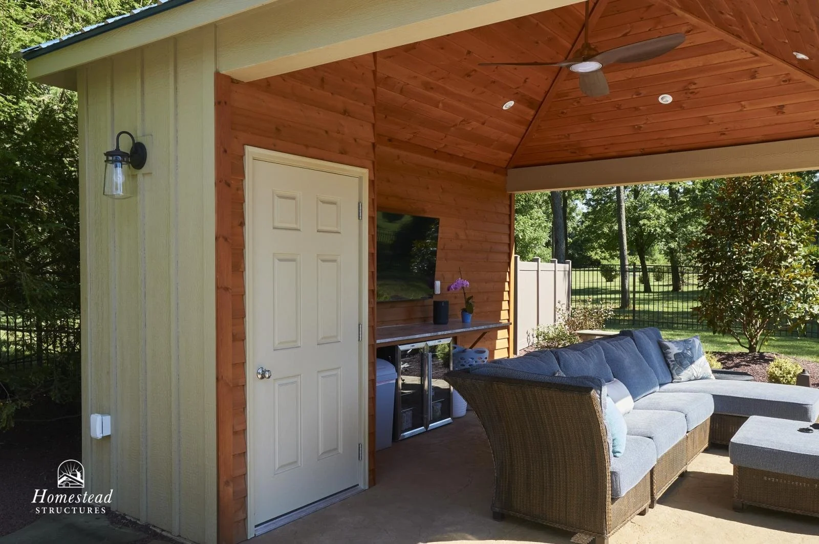 Covered outdoor patio with a wicker sectional sofa with gray cushions, a wooden table, and a TV mounted on a wood-paneled wall. There is also a modern ceiling fan and an outdoor light fixture mounted on a yellow exterior wall. The patio is surrounded
