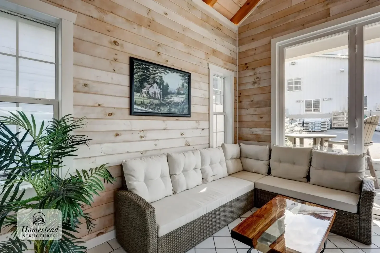 A cozy living room with a white sectional sofa, a wooden coffee table, a potted plant, a window, and a framed painting of a rural scene on a wood-paneled wall.
