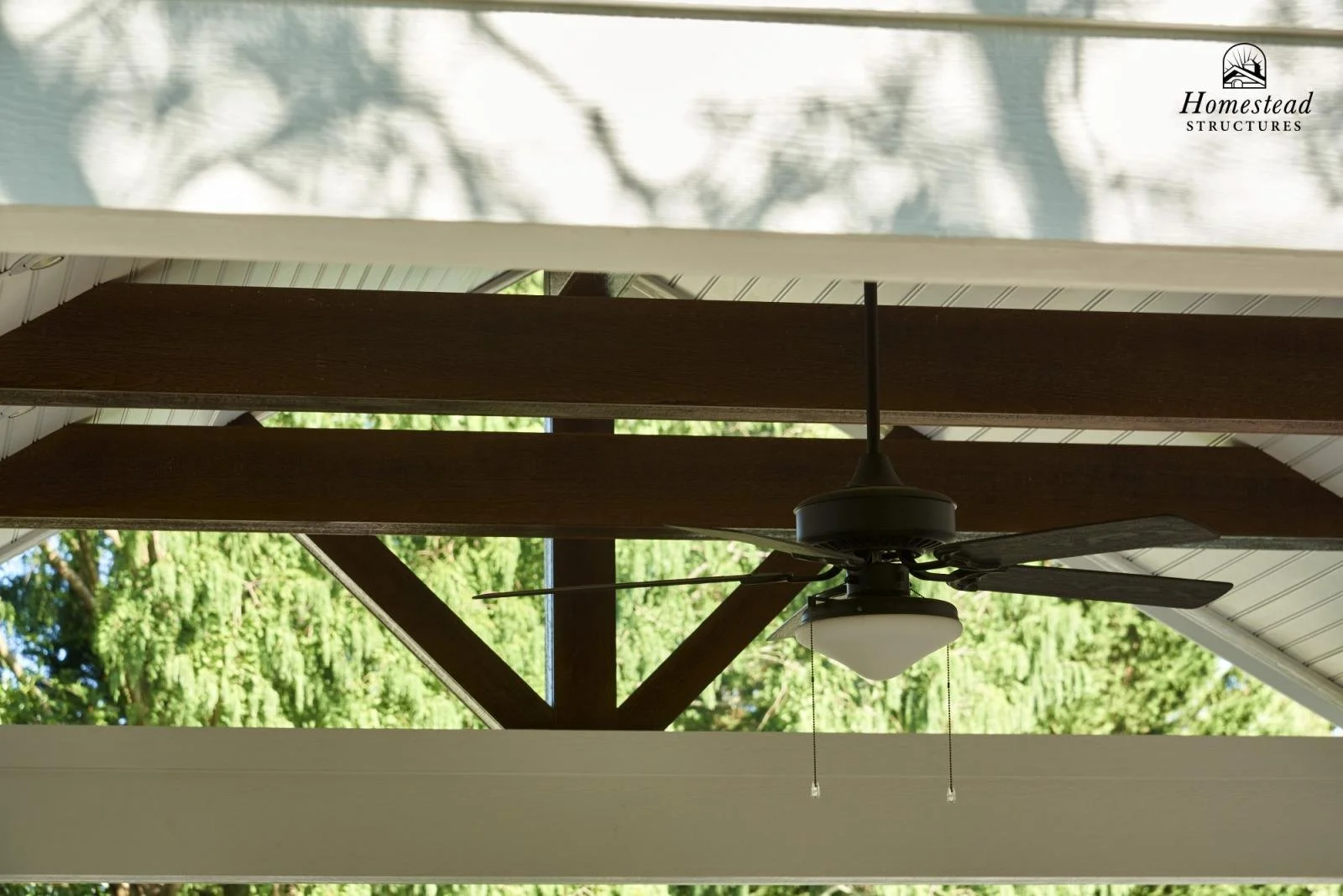 View of the underside of a house's roof with a ceiling fan and wooden beams, background shows green trees and sky.