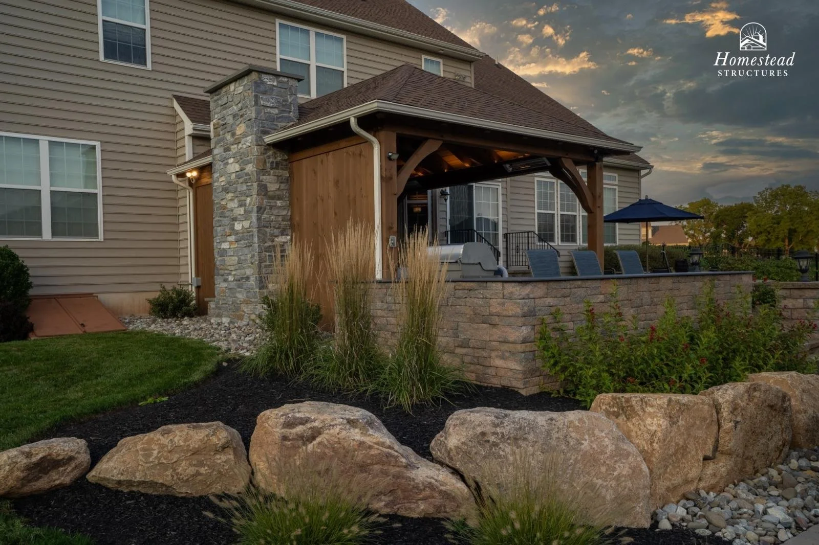 Backyard patio with stone and wood pergola, outdoor seating, umbrella, stone steps, landscaped garden with rocks, shrubs, and ornamental grass at sunset.