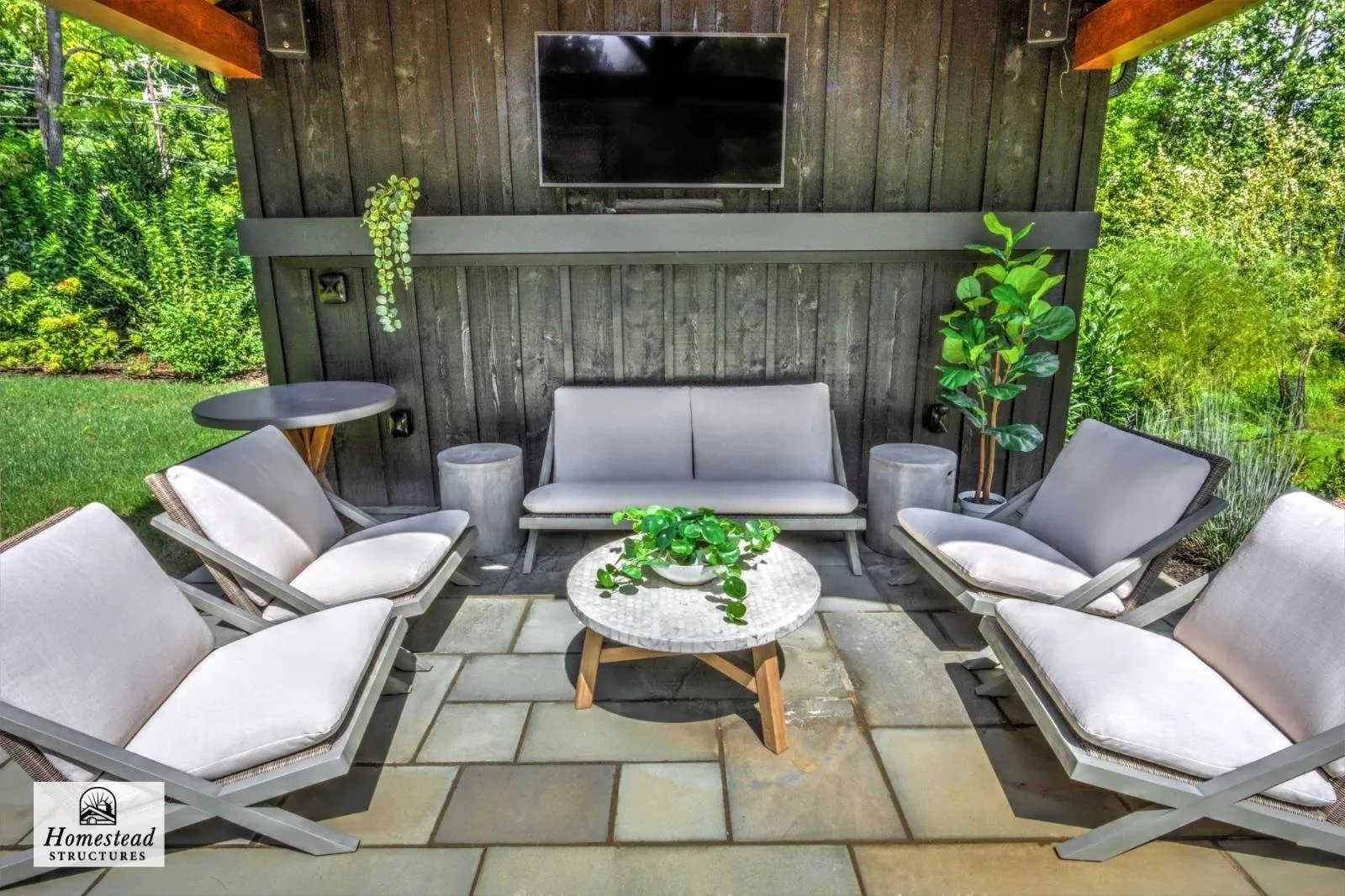 Outdoor patio seating area with six cushioned chairs, a small round table with a plant, a loveseat, a wall-mounted television, and lush greenery in the background.