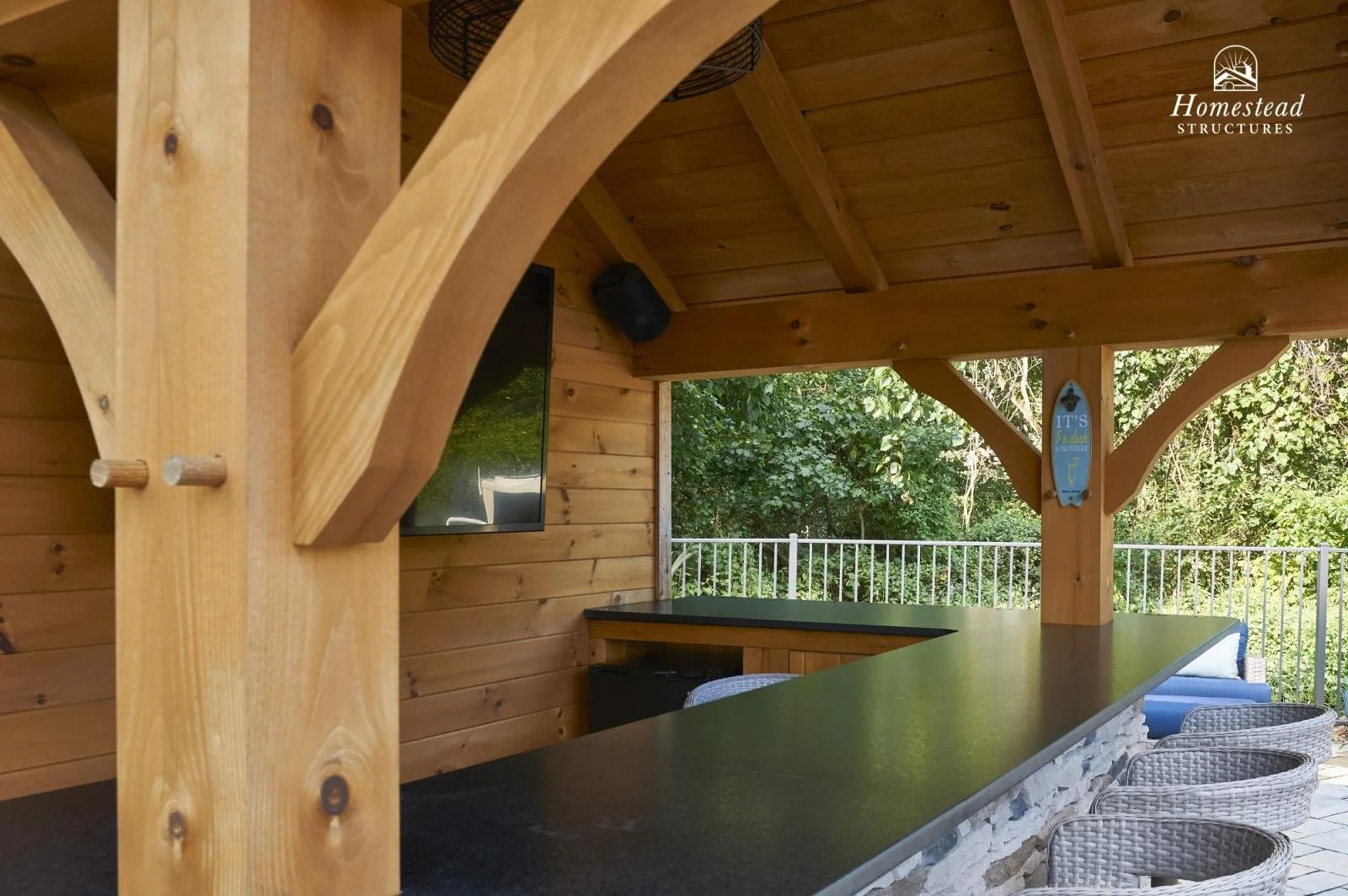 Outdoor covered patio with wooden beams, a black countertop, wicker chairs, a wall-mounted TV, and greenery in the background.