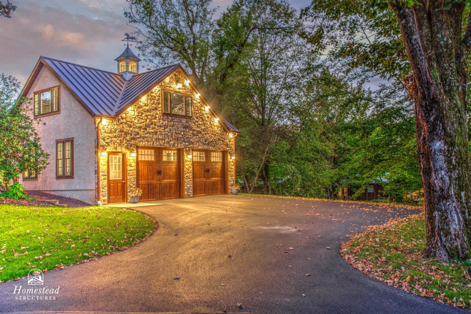 A two-story house with a stone and stucco exterior, lit with outdoor lights, surrounded by trees, with a curved driveway leading to a double garage and entrance door.