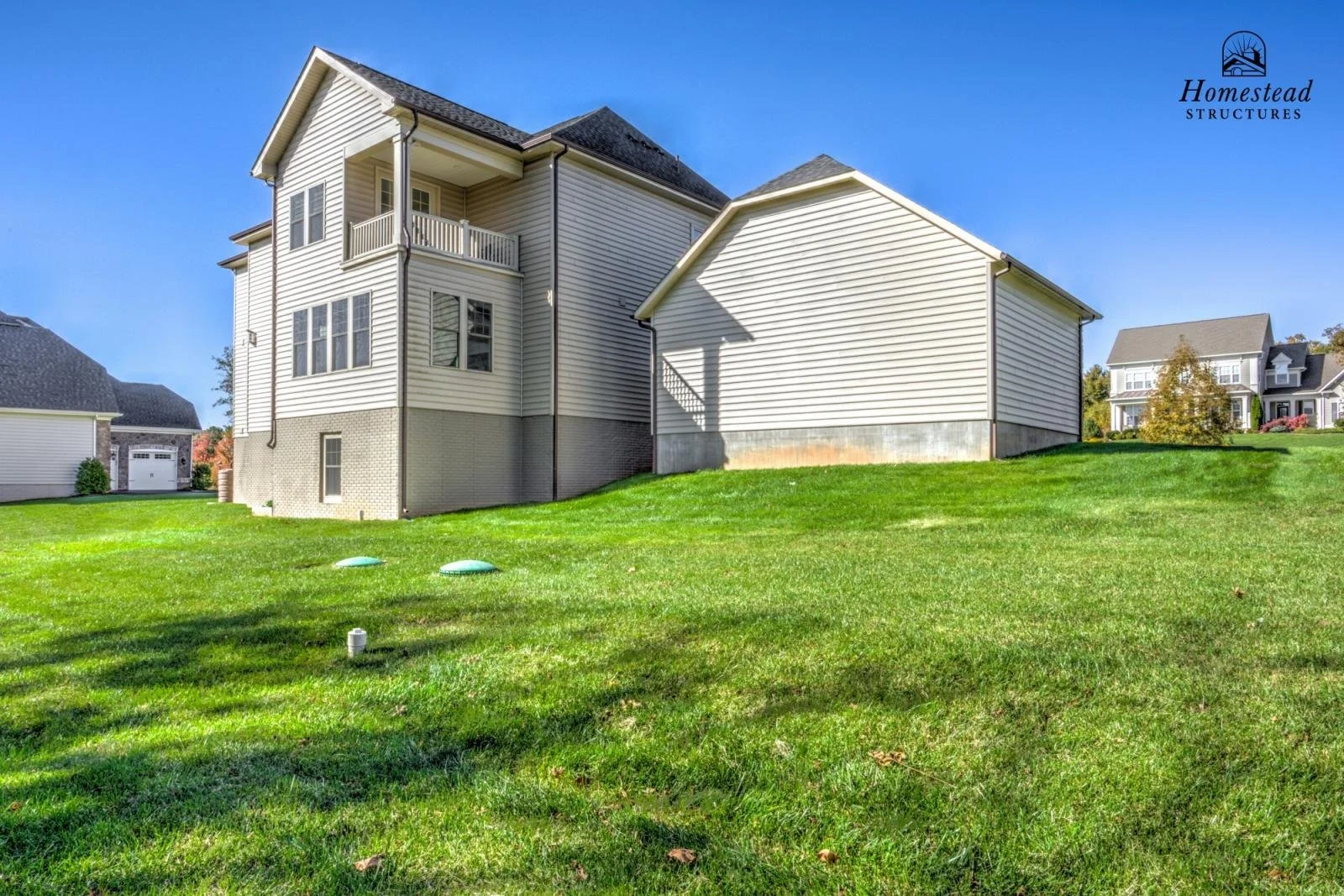 Backyard with green grass and a two-story house with beige siding and a small balcony, under a clear blue sky.