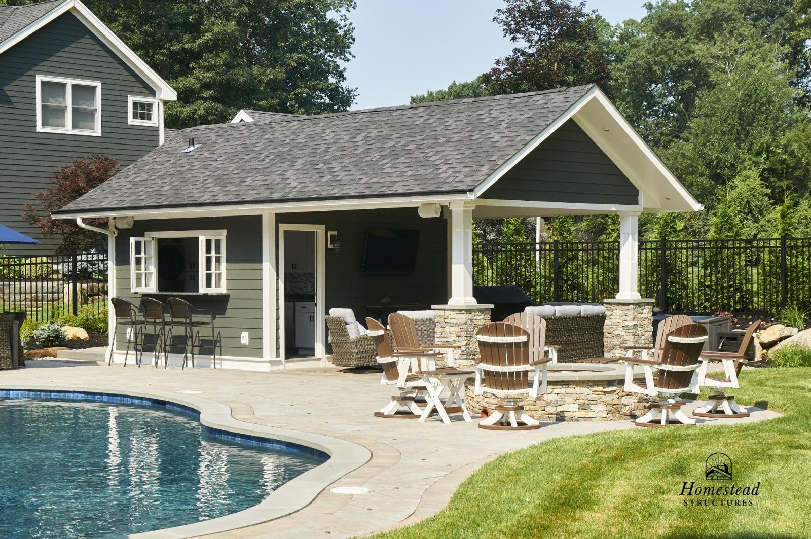 Backyard with a pool, outdoor seating area, and a pool house with a bar and television, surrounded by a black fence and green trees.