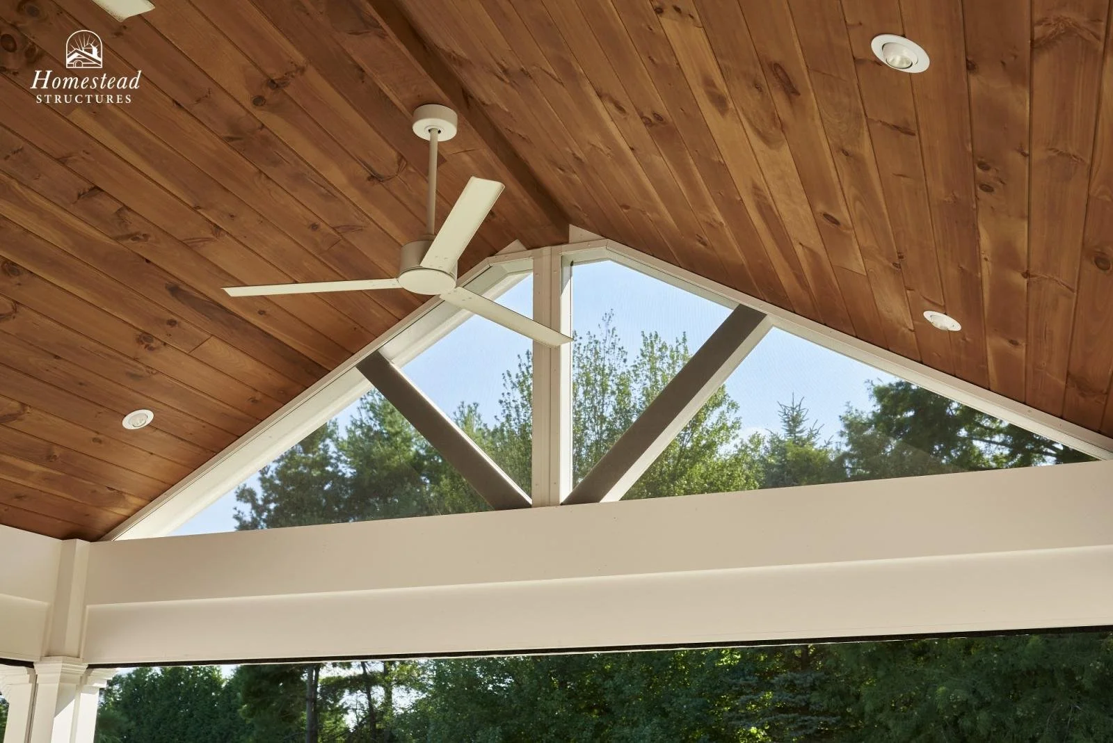 Ceiling with wooden panels, white ceiling fan, triangular window revealing trees outside, and recessed lighting