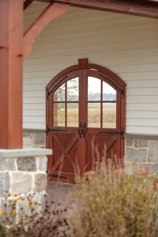 Wooden barn door with glass panes on a house with white siding and stone foundation, viewed through some plants.