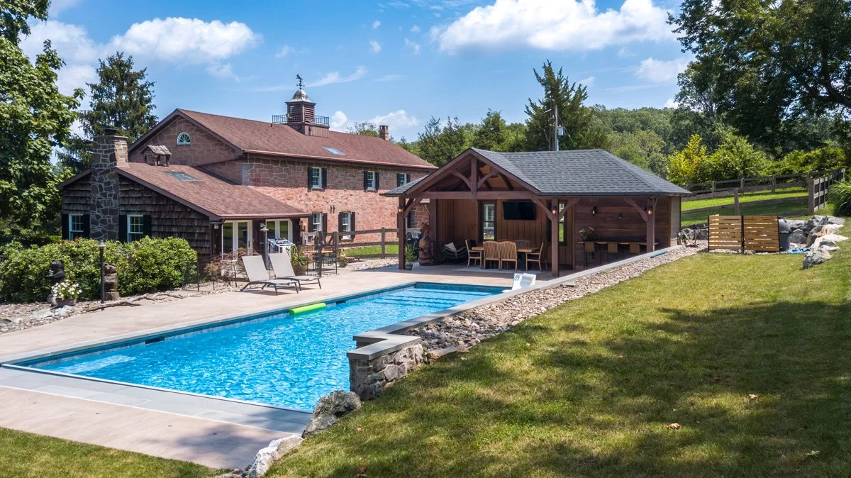 A backyard with a swimming pool, a paved deck with lounge chairs, a wooden pool house with outdoor furniture, and a large house in the background surrounded by trees and green grass.