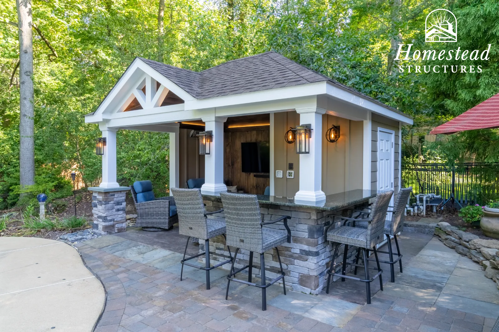 An outdoor patio area featuring a small bar with woven bar stools, a covered pavilion with outdoor seating, surrounded by lush green trees and landscaping.