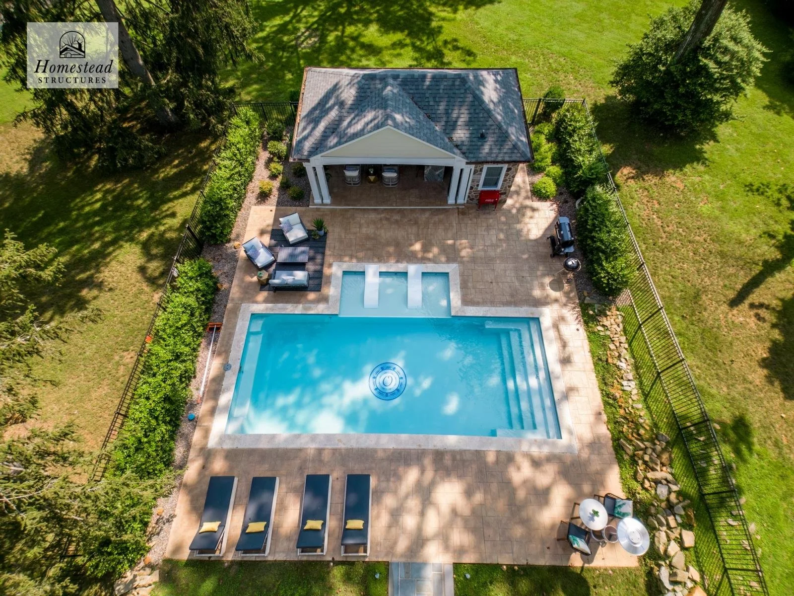 Aerial view of a backyard with a rectangular swimming pool, surrounded by a tan tile deck, with seating areas and a small covered patio. The yard is enclosed with a black metal fence and has greenery including trees and bushes.