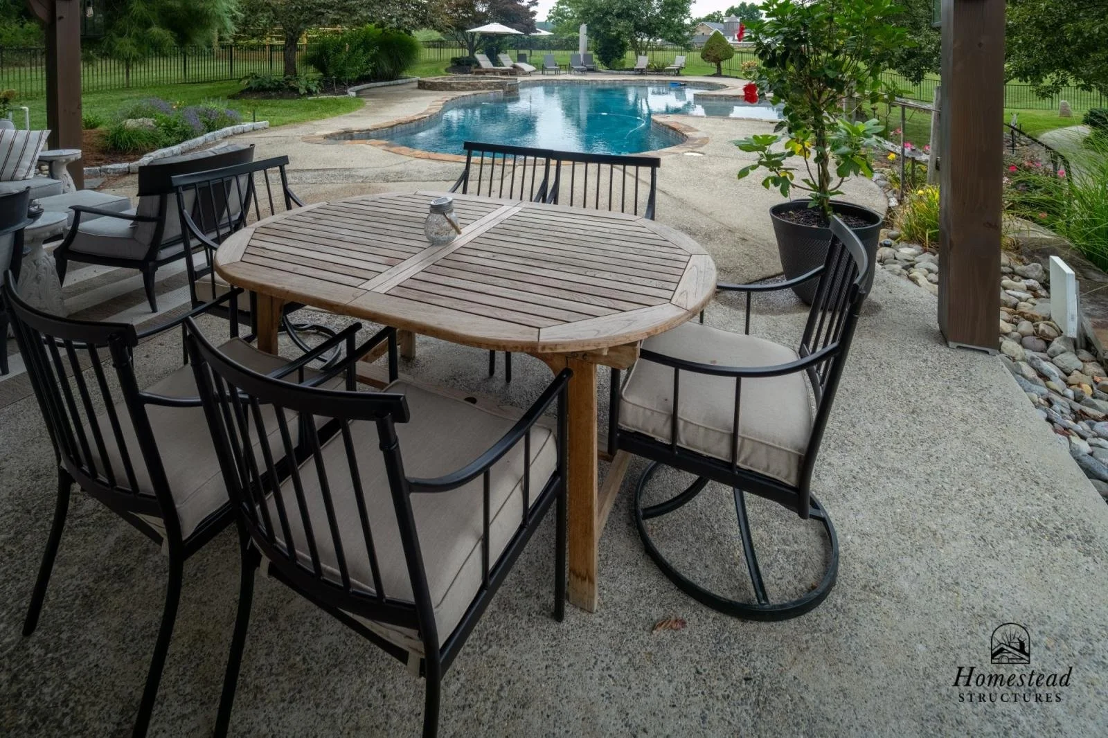 Outdoor patio with a wooden round table and black chairs, overlooking a swimming pool and landscaped backyard.