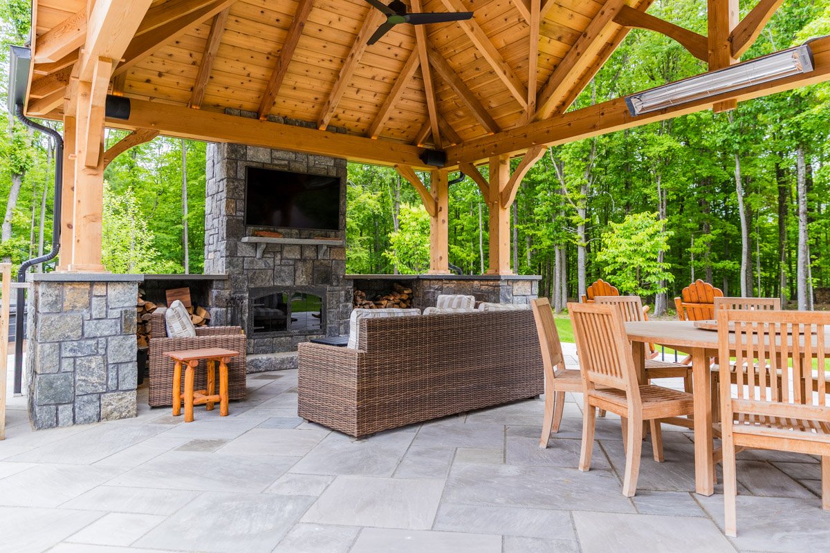 Outdoor covered patio with wooden roof, stone fireplace, outdoor furniture including wicker and wooden chairs and a large dining table, surrounded by green trees.