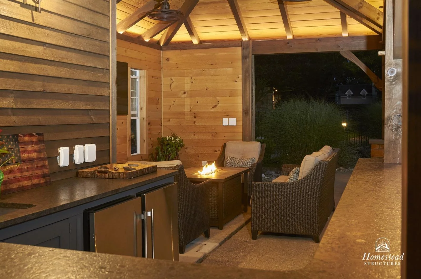 Outdoor patio area with a fire pit surrounded by wicker chairs, a counter with a cutting board, and a wooden wall with a small window, at night.