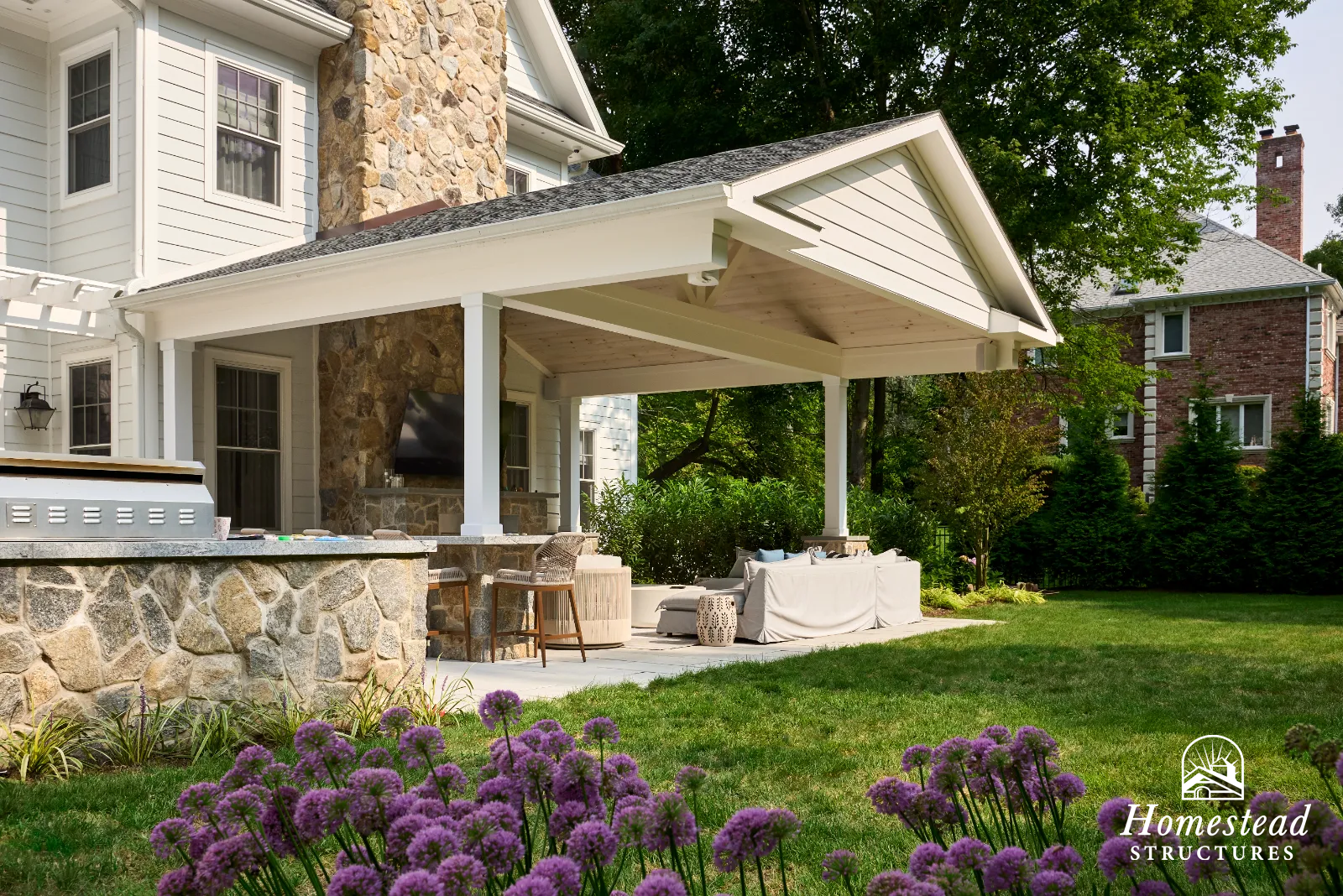 Backyard patio area with covered porch, outdoor seating, stone accents, and surrounding greenery, with a lawn and flowers in the foreground.