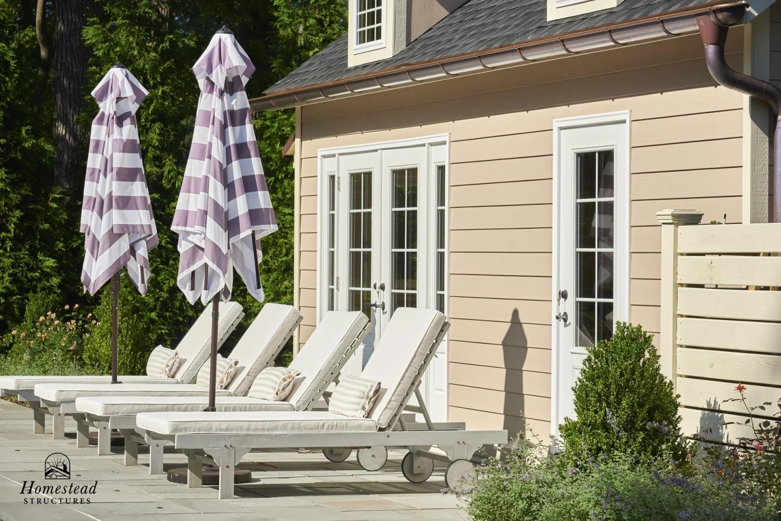 A backyard patio with three white lounge chairs with striped pillows, purple and white striped umbrellas, a beige house with white trim, and some greenery and plants.