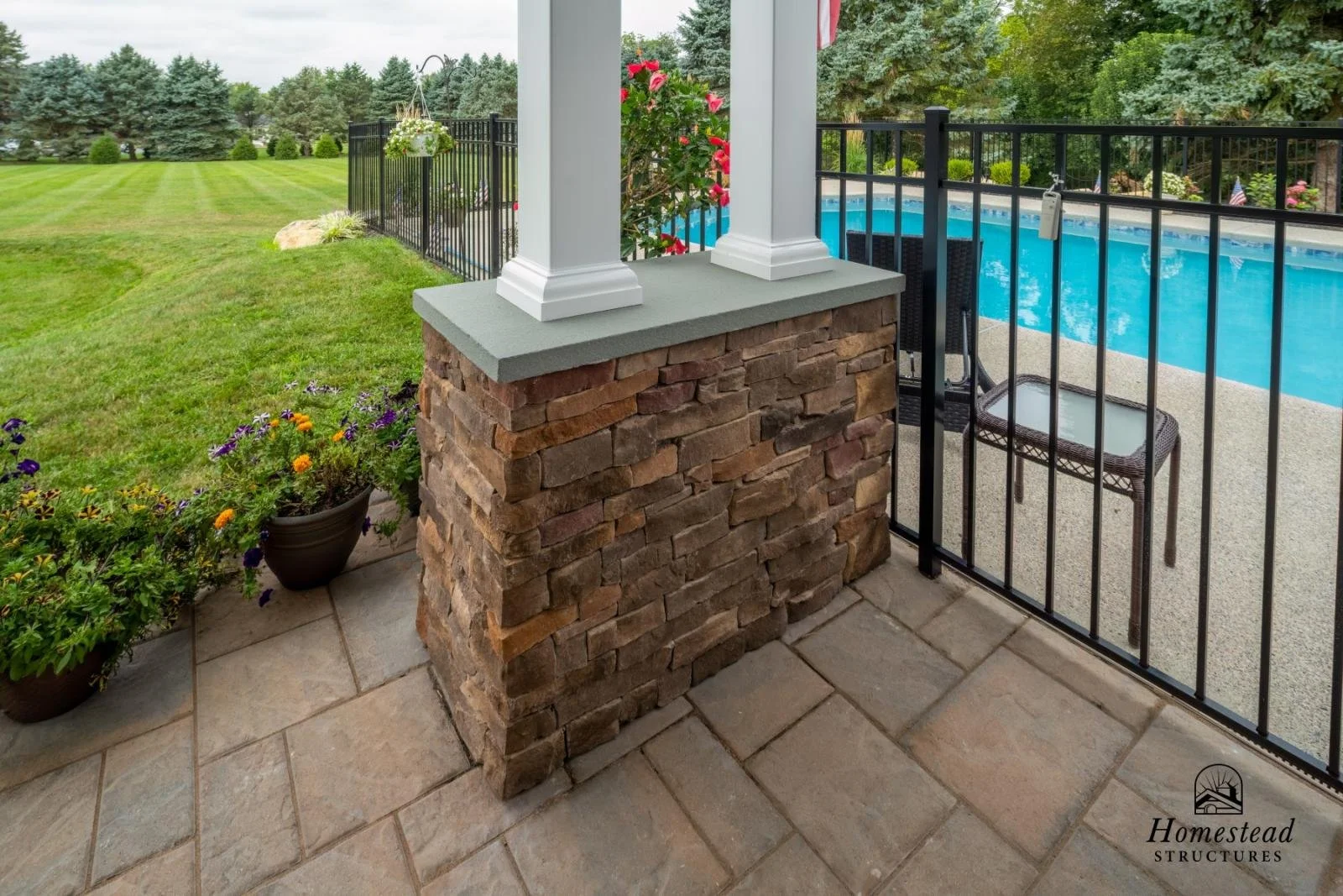 Close-up of a covered patio with a brick pillar, potted flowers, and a view of a backyard pool surrounded by a black metal fence, with a lush green lawn and trees in the background.