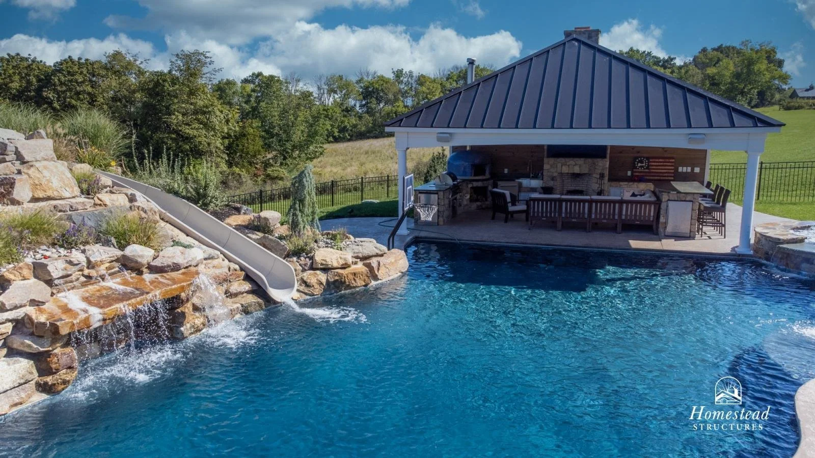 Backyard swimming pool with waterfall, slide, stone waterfall feature, covered outdoor kitchen and lounge area, surrounded by greenery and fencing.