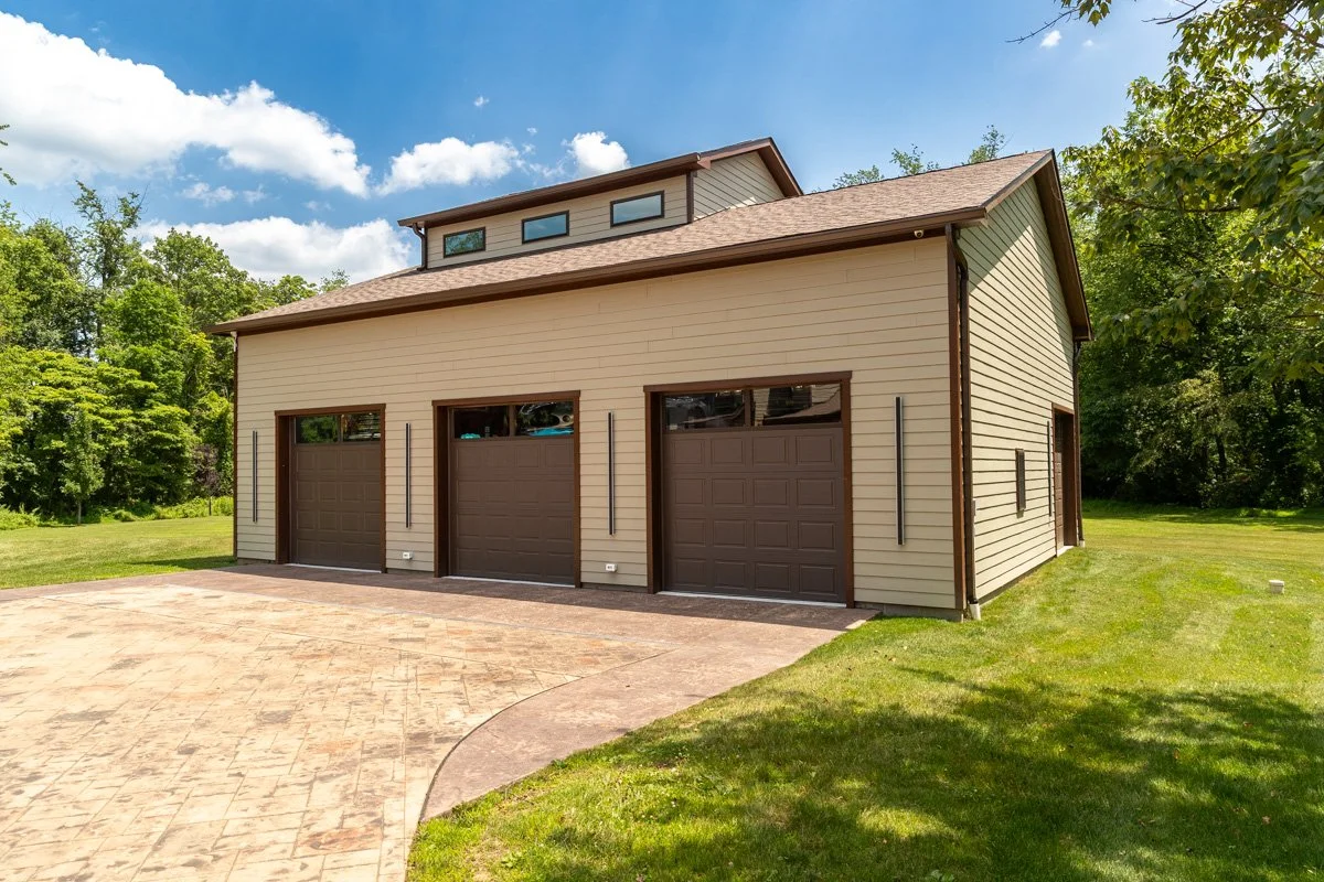 A modern beige garage building with three garage doors and a driveway, surrounded by green grass and trees, under a blue sky with some clouds.
