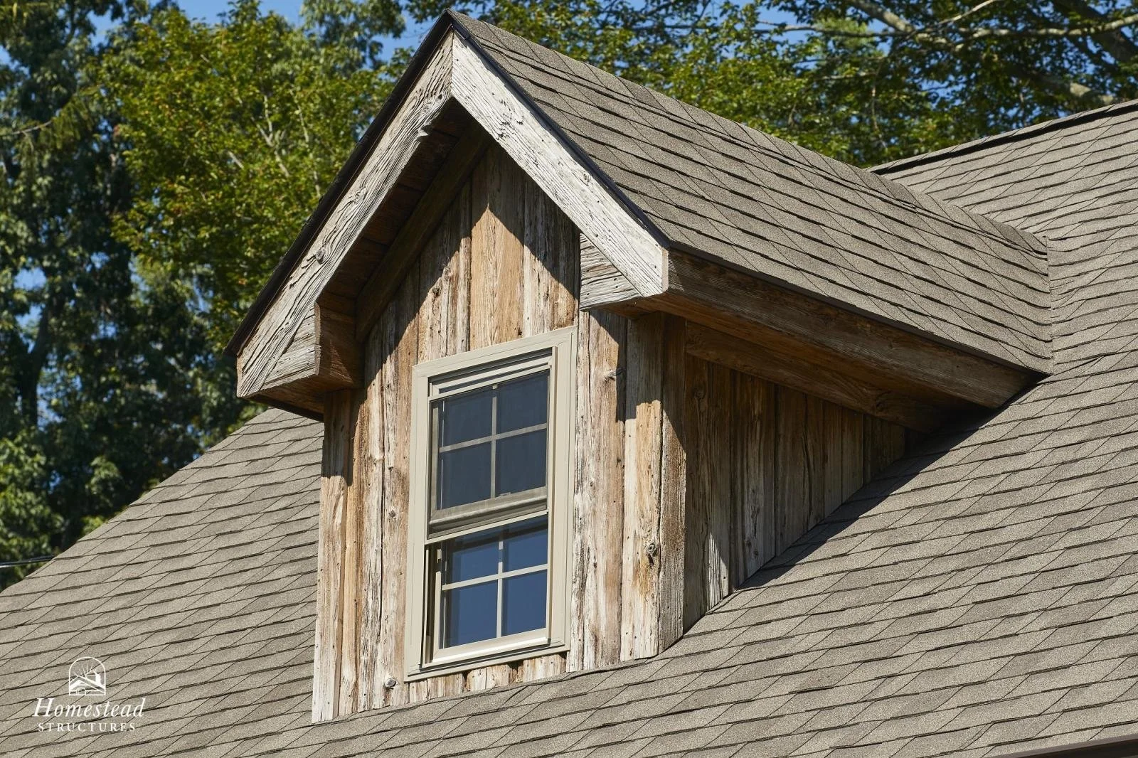 Close-up of a dormer window on a house with a shingled roof and weathered wooden siding, surrounded by green trees.