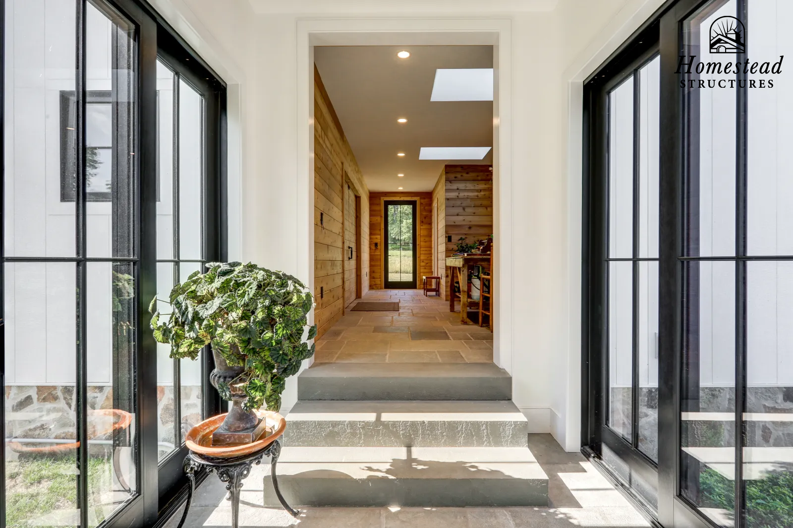 View from a porch into a hallway with wooden walls and a door at the end, featuring black-framed glass doors and a potted plant on a decorative table.
