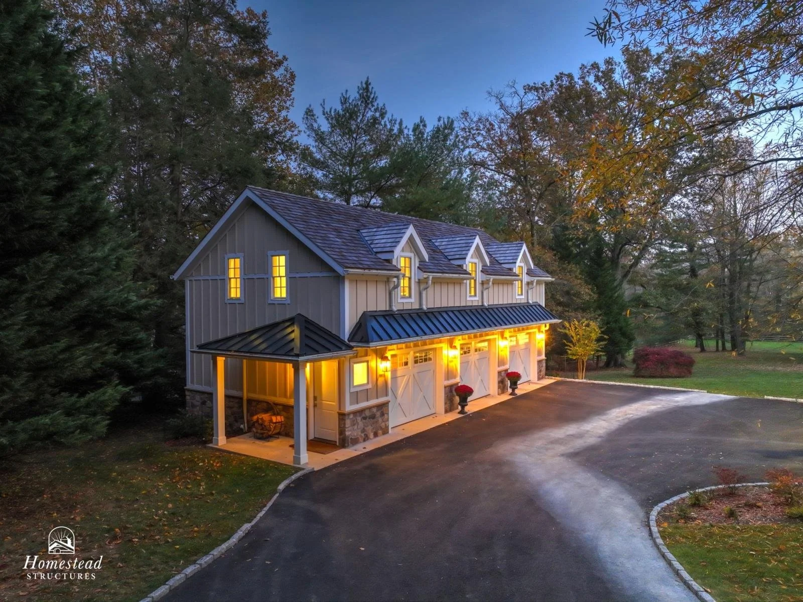 A two-story garage with a gray exterior, white trim, and a dark metal roof, illuminated from within, surrounded by trees with fall foliage.