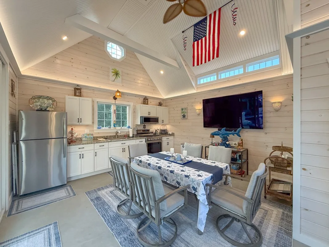 Open kitchen and dining area with white cabinets, stainless steel refrigerator, and a wall-mounted TV. Features a table with a floral tablecloth and six chairs, a window over the sink, and patriotic decorations including an American flag hanging from
