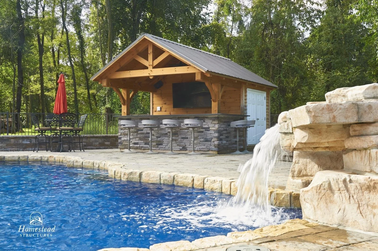 Outdoor swimming pool with a waterfall feature, poolside seating, and a wooden pool house with a stone base in a wooded setting.