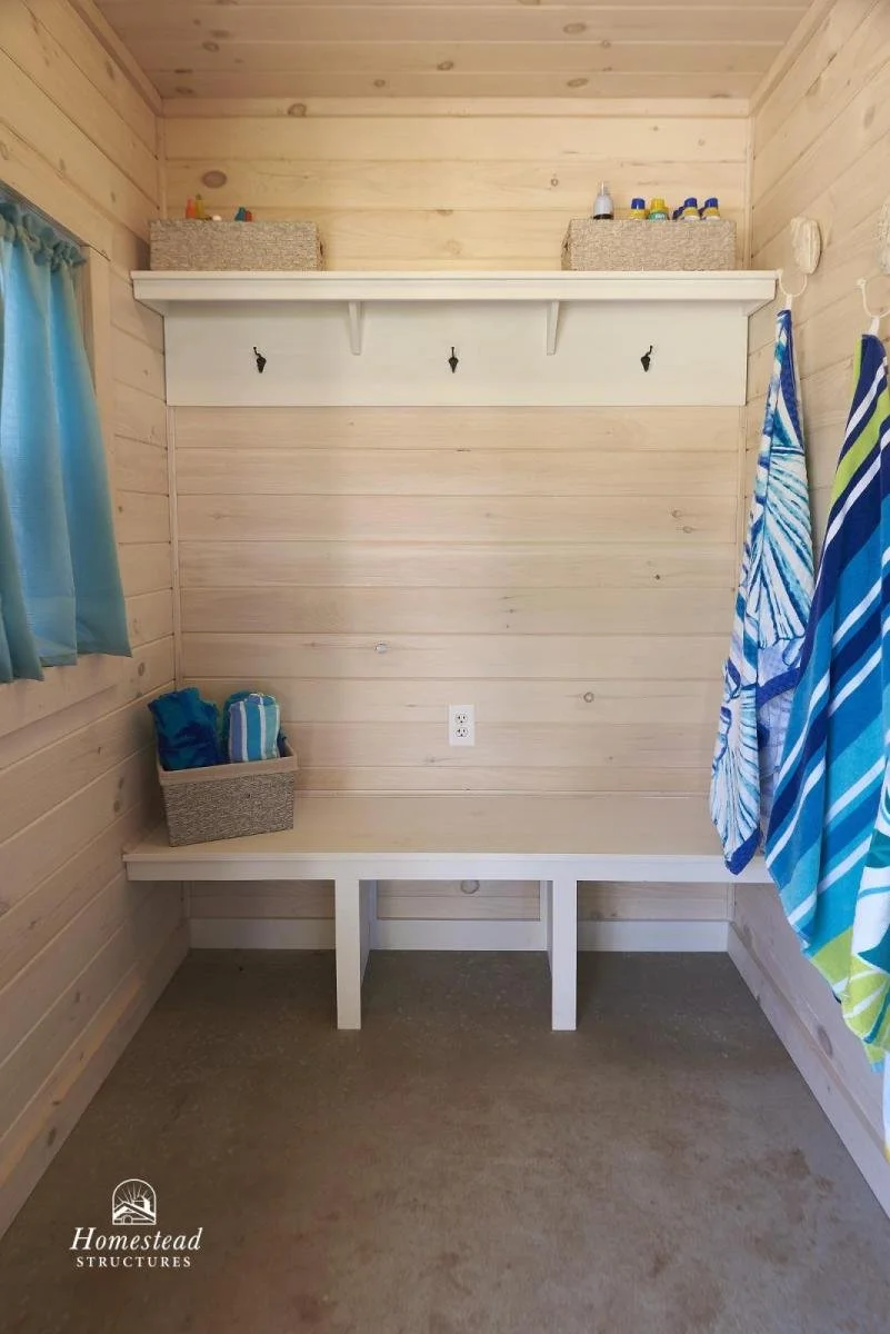 An empty wooden room with a built-in bench, hanging hooks, a shelf with storage baskets, and towels hanging on hooks, likely part of a beach or laundry area.