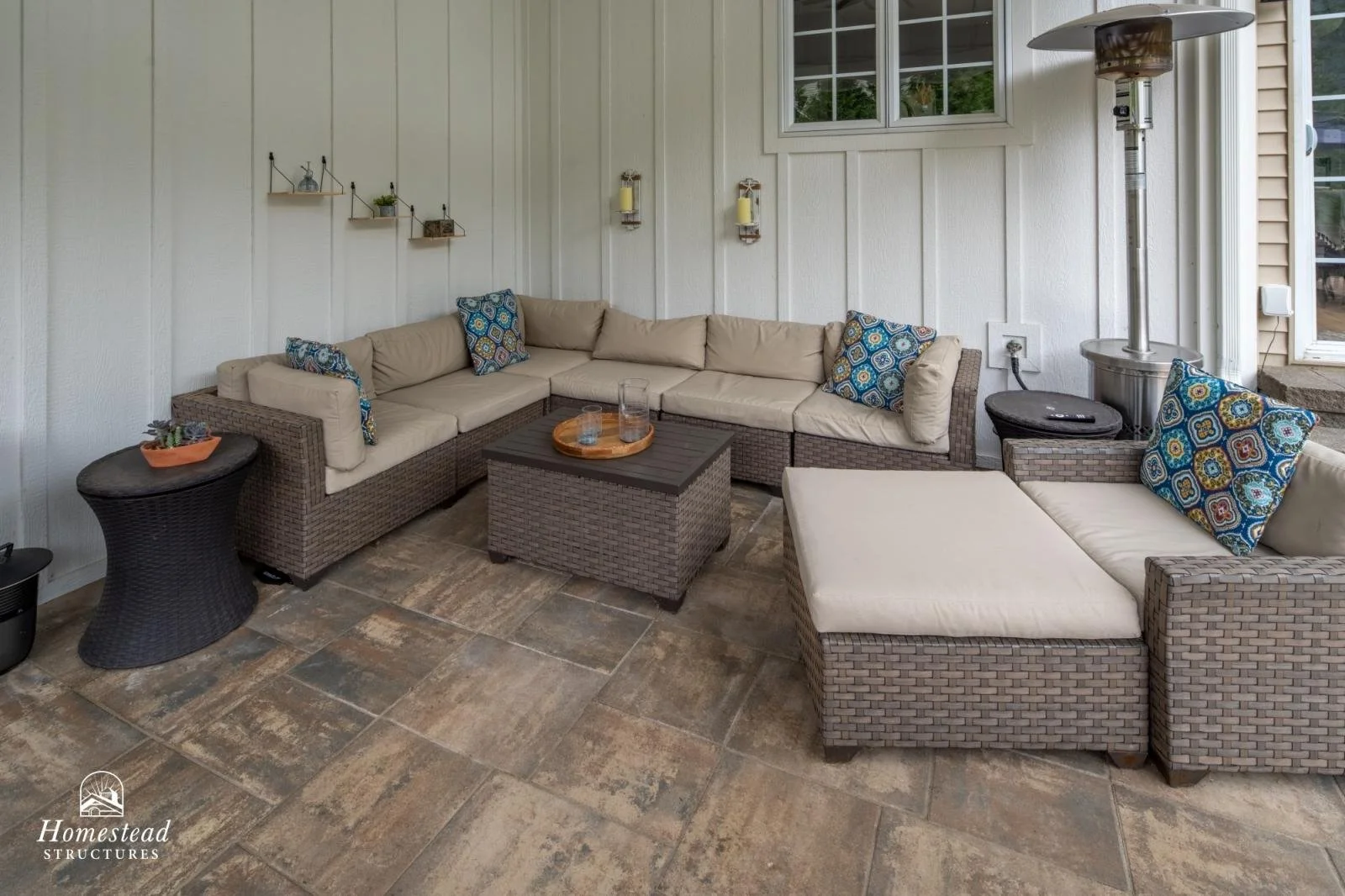 Covered outdoor patio with a large L-shaped wicker sectional sofa with beige cushions and decorative pillows, a matching wicker ottoman, small side tables, a patio heater, and wall-mounted candle holders, with a window and siding on the wall.