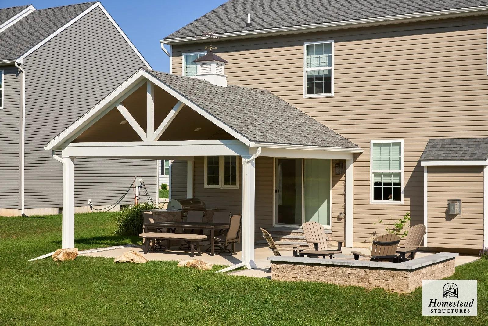 Backyard patio with outdoor furniture under a covered pergola attached to a beige two-story house with white trim and multiple windows. The patio includes a dining table, chairs, Adirondack chairs, and a built-in brick firepit area, with a green lawn
