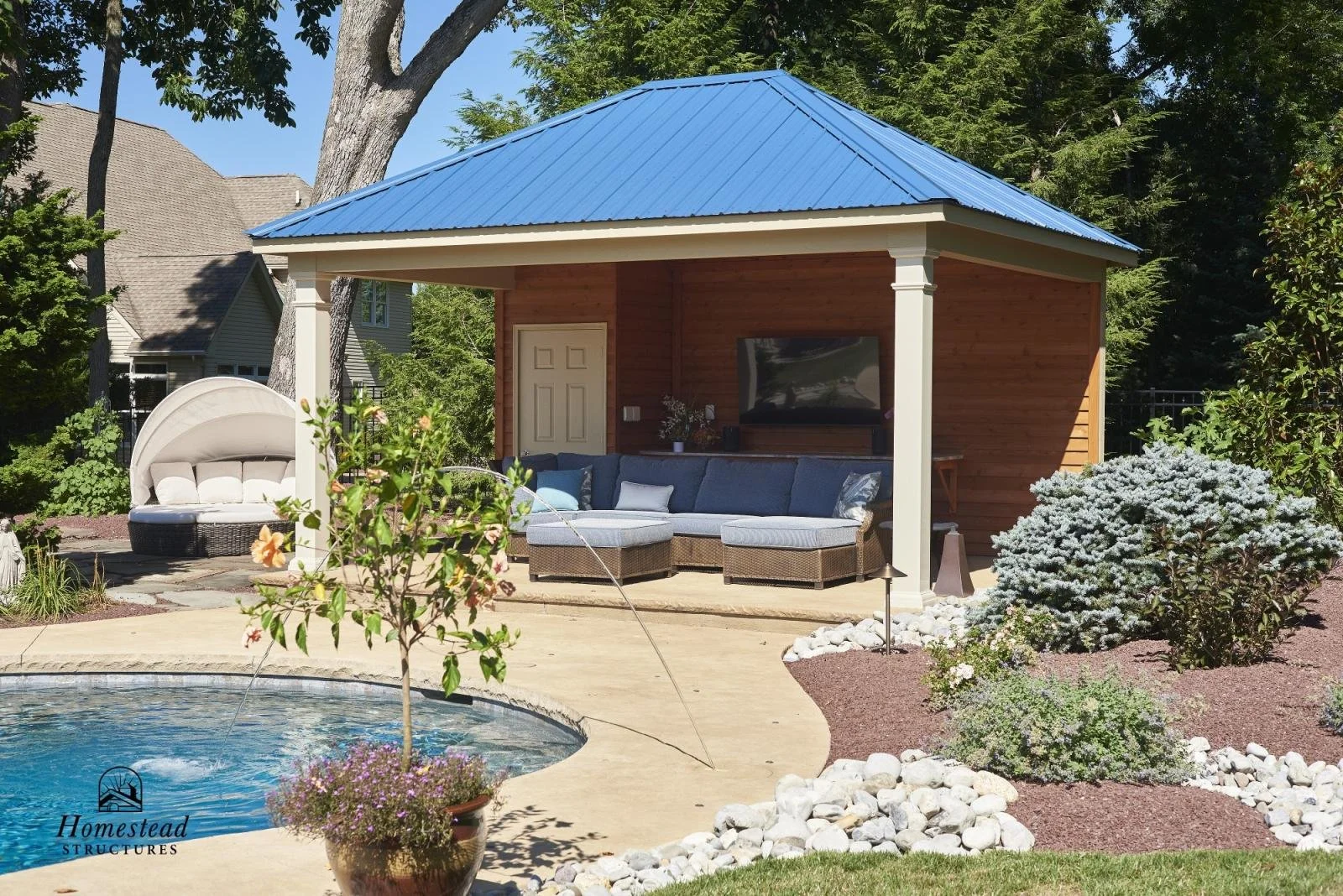 Backyard patio with a small pool, potted plant, seating area with outdoor furniture, and a wooden structure with a blue metal roof surrounded by greenery.