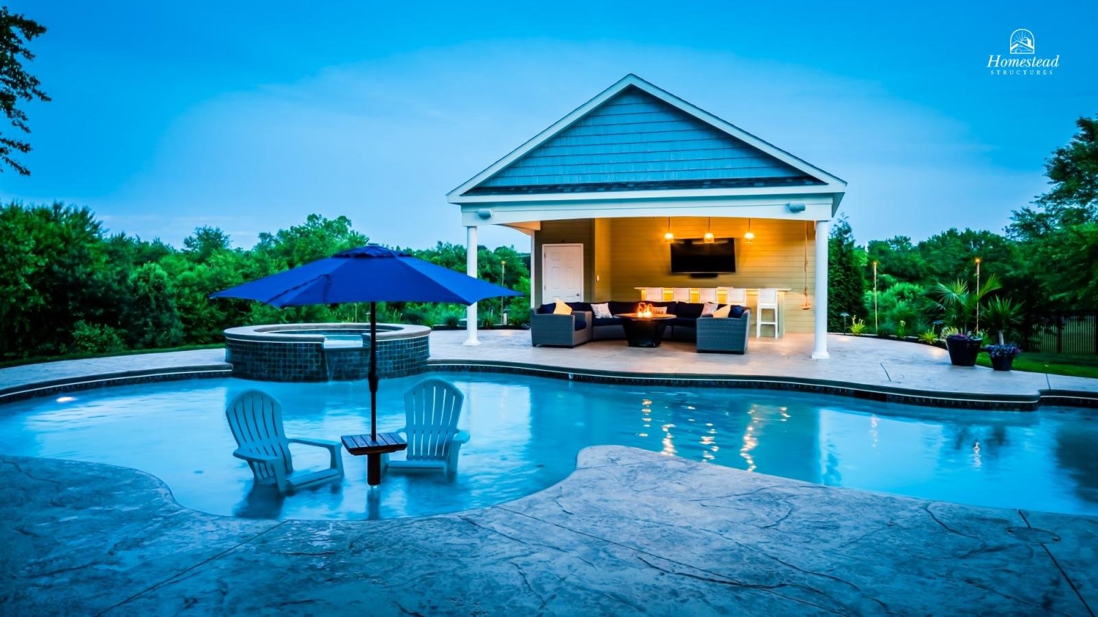 A backyard with a swimming pool, two white chairs under a blue umbrella, a hot tub on the pool deck, and a covered outdoor living area with a sofa, fire pit, and TV, surrounded by green trees.