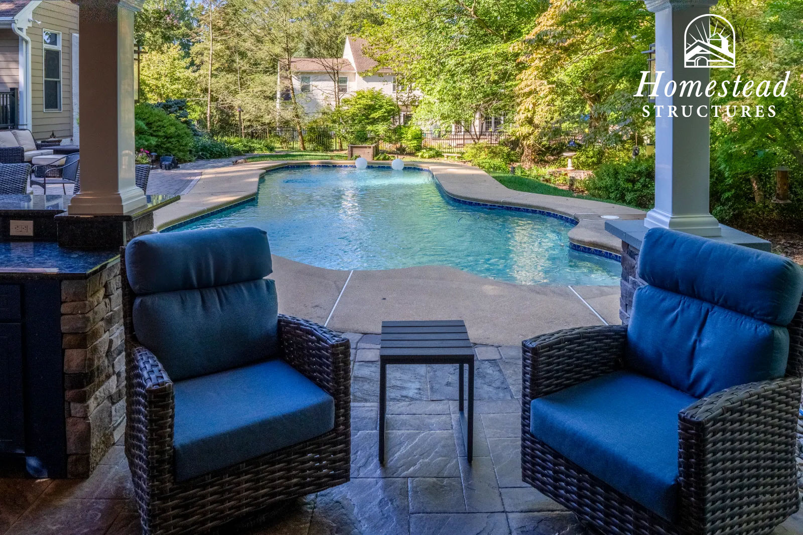 Porch with two wicker chairs with blue cushions facing a swimming pool surrounded by trees and a backyard with additional seating and a fenced yard.