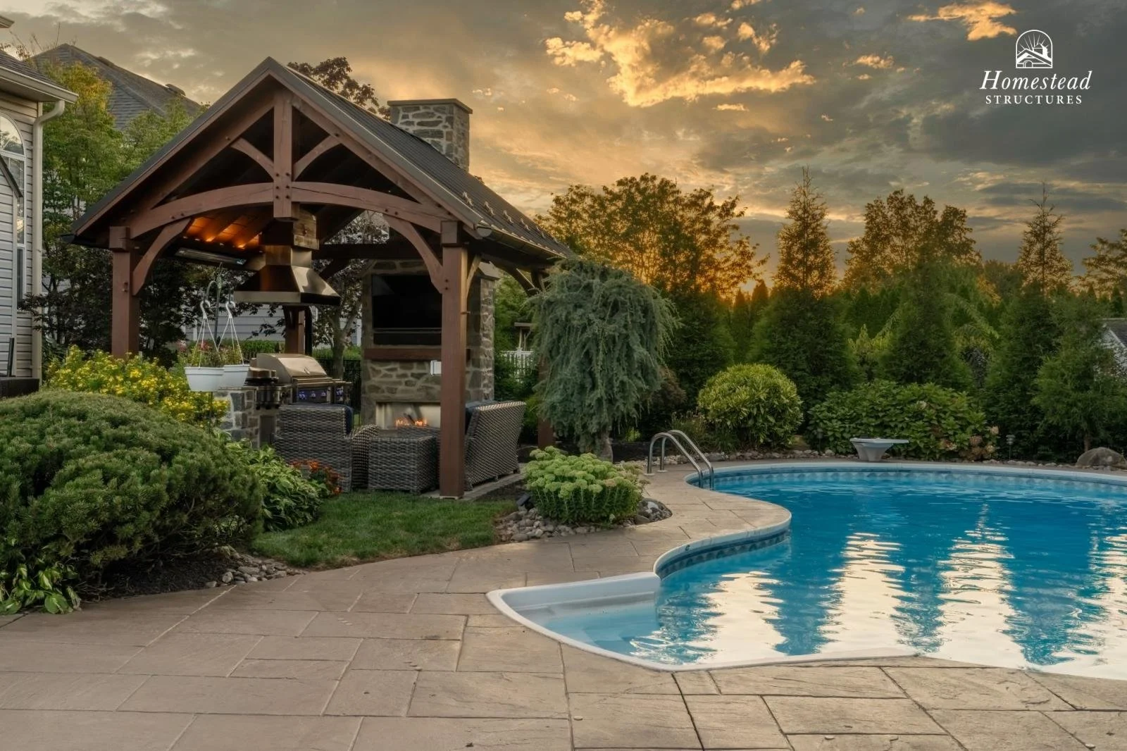 Backyard with a swimming pool, a fire pit with outdoor seating, a covered outdoor kitchen with a grill, and surrounded by trees and landscaping at sunset.