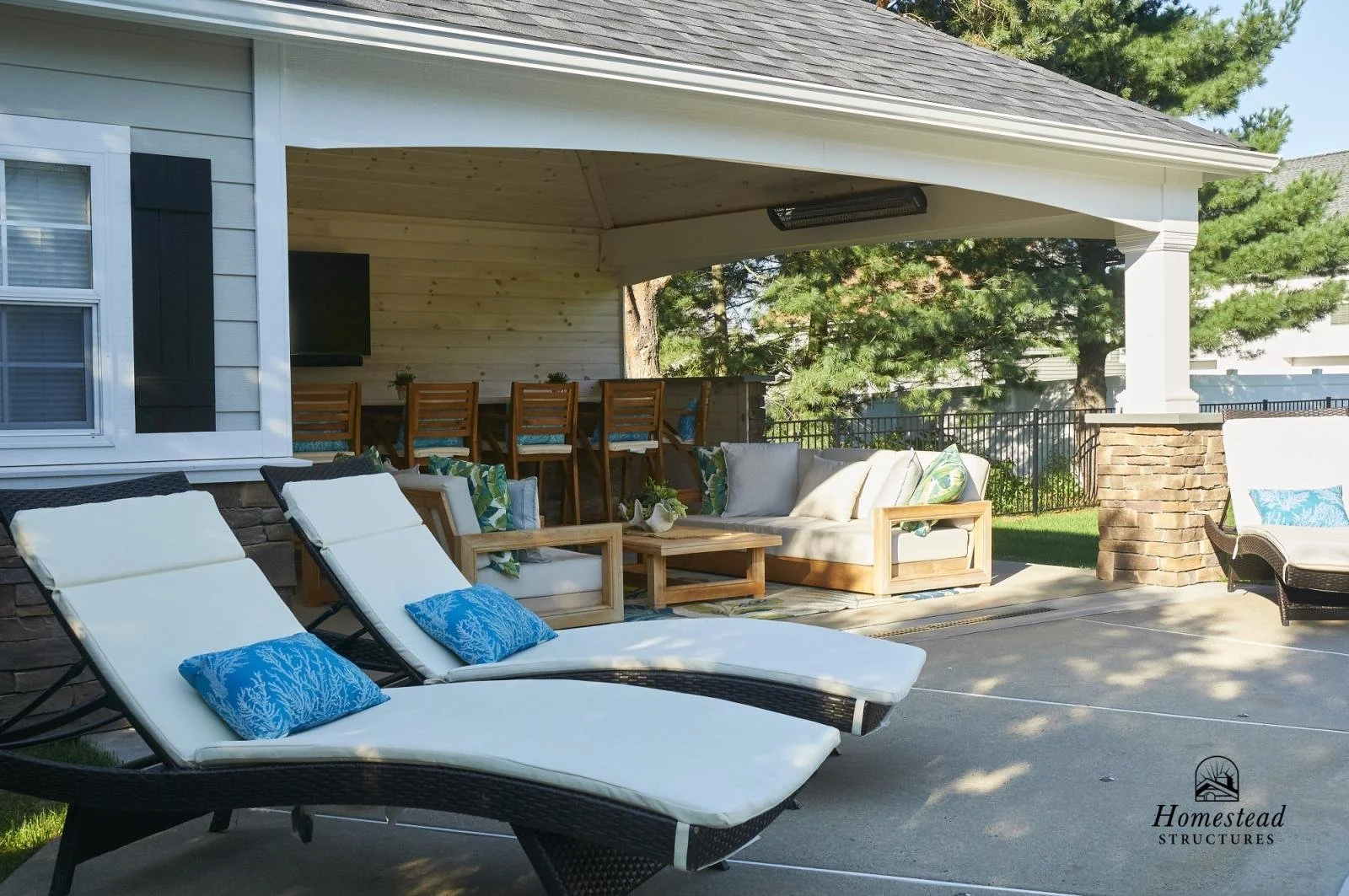 A backyard patio with two white lounge chairs with blue pillows, a wooden sofa with white cushions and patterned pillows, and a wooden bar with barstools in front of a house with white siding and black shutters. There are trees and a grassy yard beyo